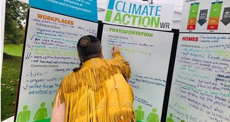 A person writing on a large presentation board. The question on the board is: What should be in place in Waterloo Region to make it possible for you to reduce your emissions at work, home and in transportation?