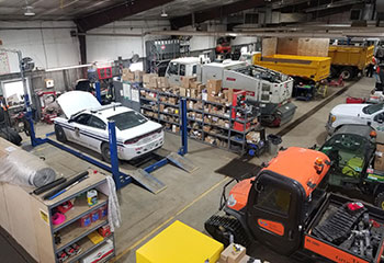A number of vehicles and other equipment parked inside a large garage-type building, with one car’s hood up for maintenance.