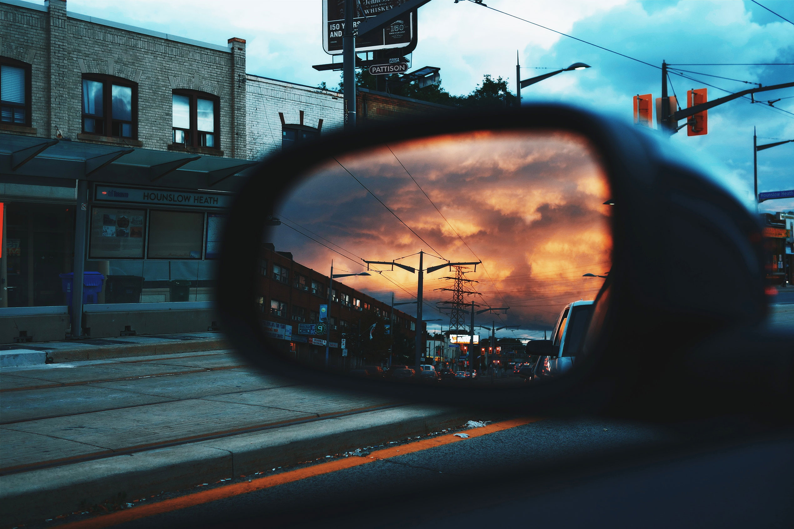 Photo showing the driver's side car mirror reflecting an orange sunset, with the car visibly traveling down a city street with distant street lights.