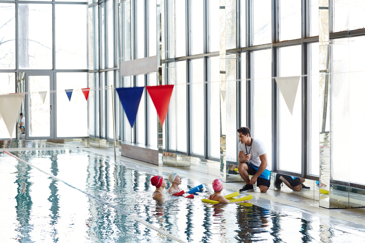Big windows around an indoor swimming pool, an instructor leaning down talking to a few swimmers