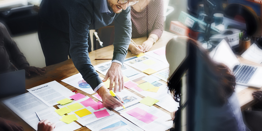 a group of people collaborating around a table filled with documents, laptops, and sticky notes, engaged in a planning or brainstorming session led by one individual.