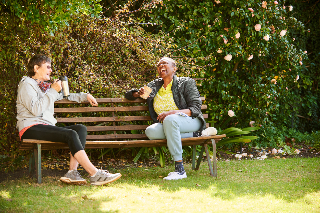 Two friends sitting on bench together and laughing in sunny park.