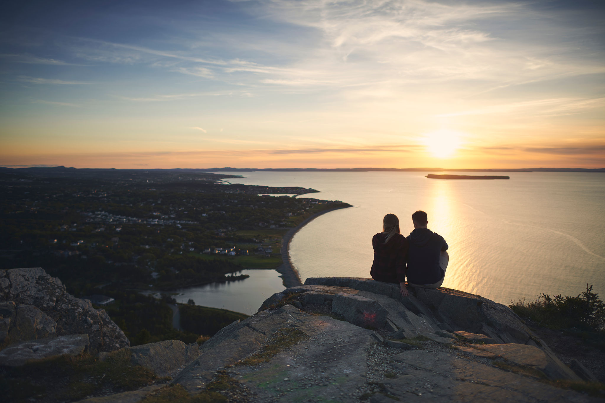 Two people sitting high on a bluff, looking out at the coastline as the sun sets over the ocean.