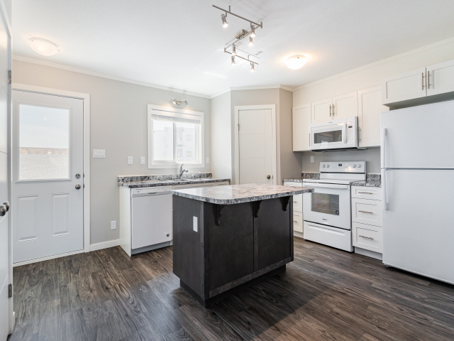 L'intérieur d'une maison dans le lotissement Aspen Heights à Saskatoon. L'image montre une cuisine toute neuve équipée d'un réfrigérateur, d'une cuisinière, d'un îlot et d'un comptoir. Les planchers sont en bois foncé et les armoires et appareils électroménagers sont blancs.