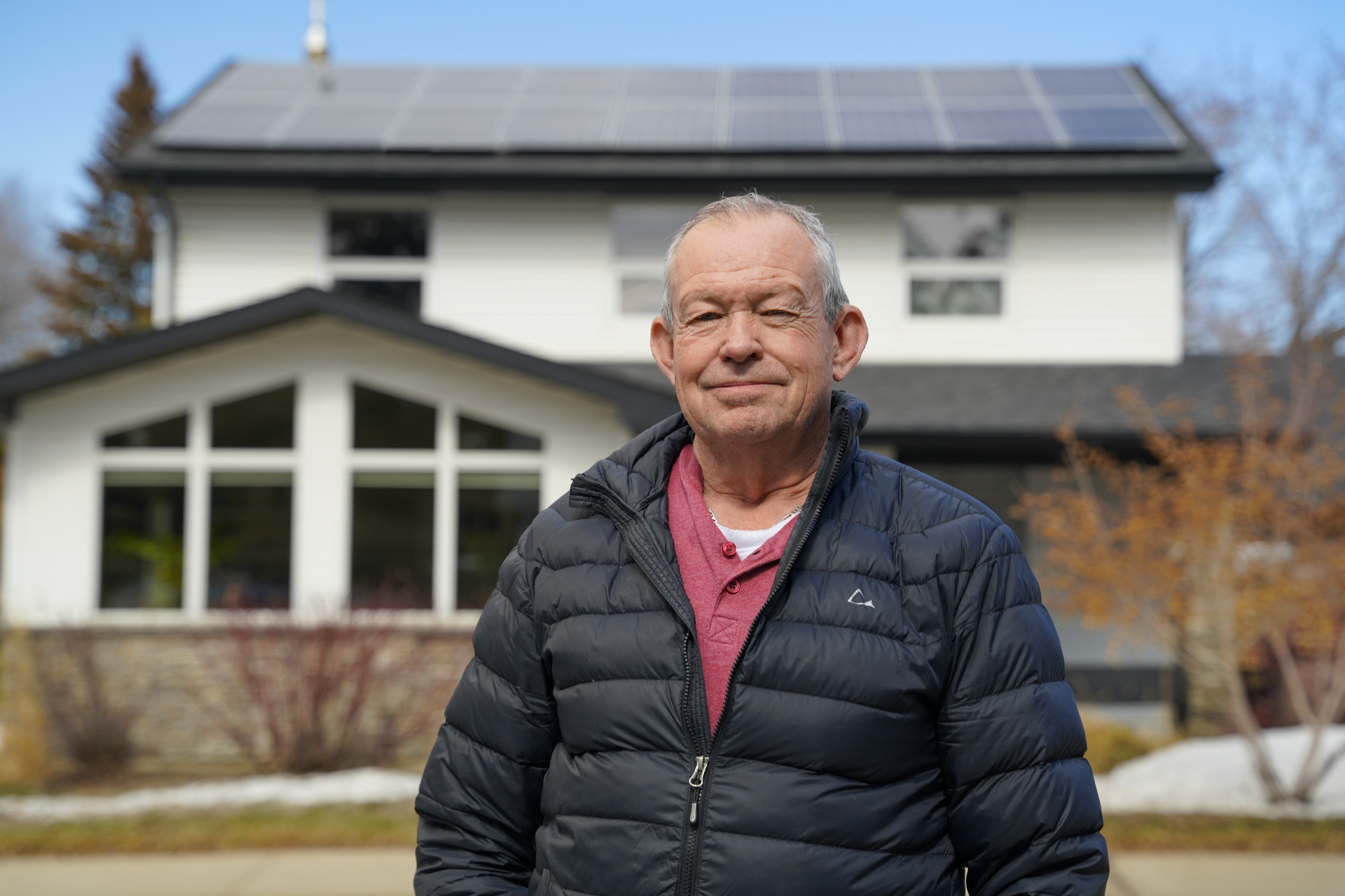 A person wearing a black puffy jacket standing in front of a white house with solar panels on its roof.