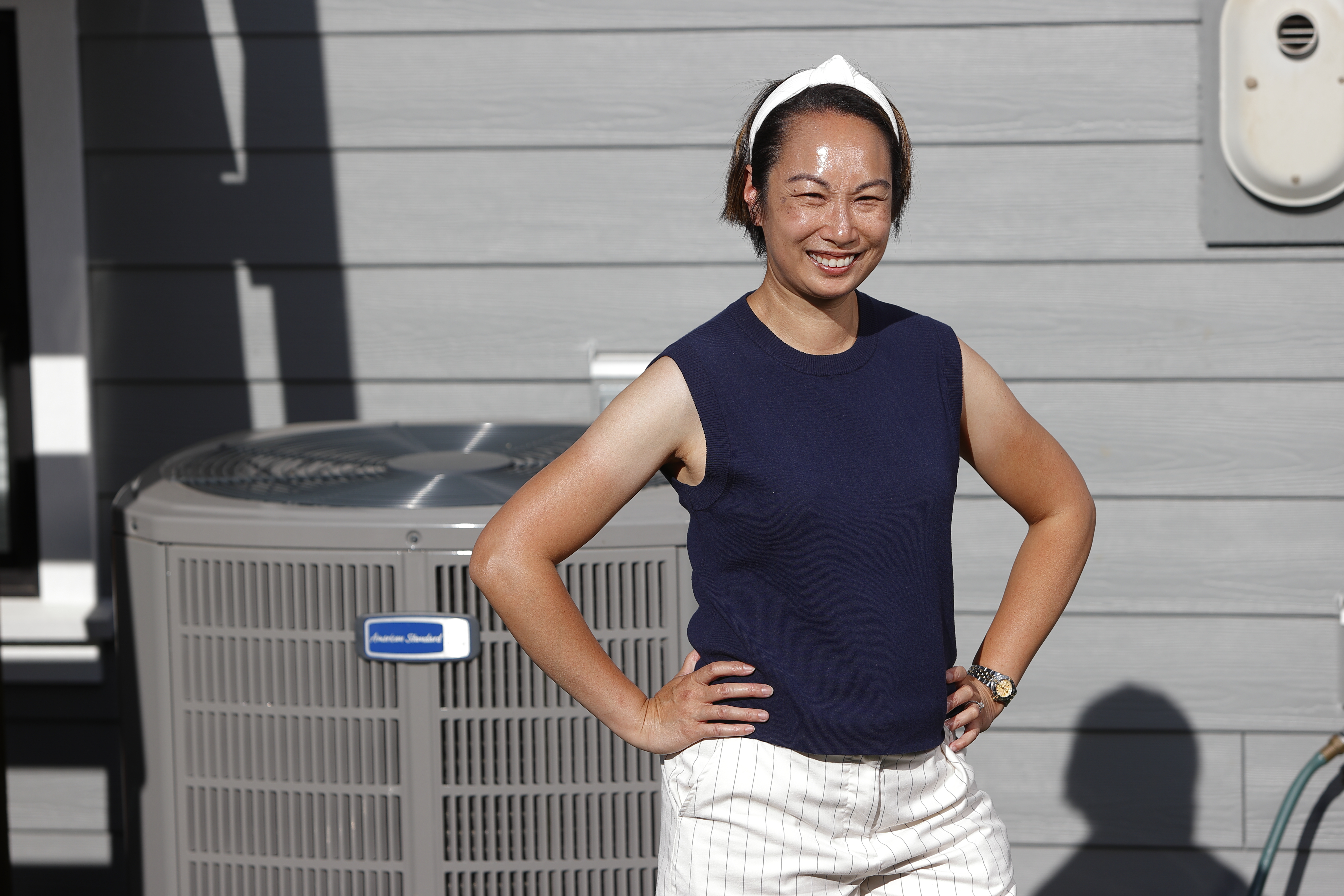 A person with hands on hips stands outside their home, in front of an outdoor heat pump unit, on a sunny day.