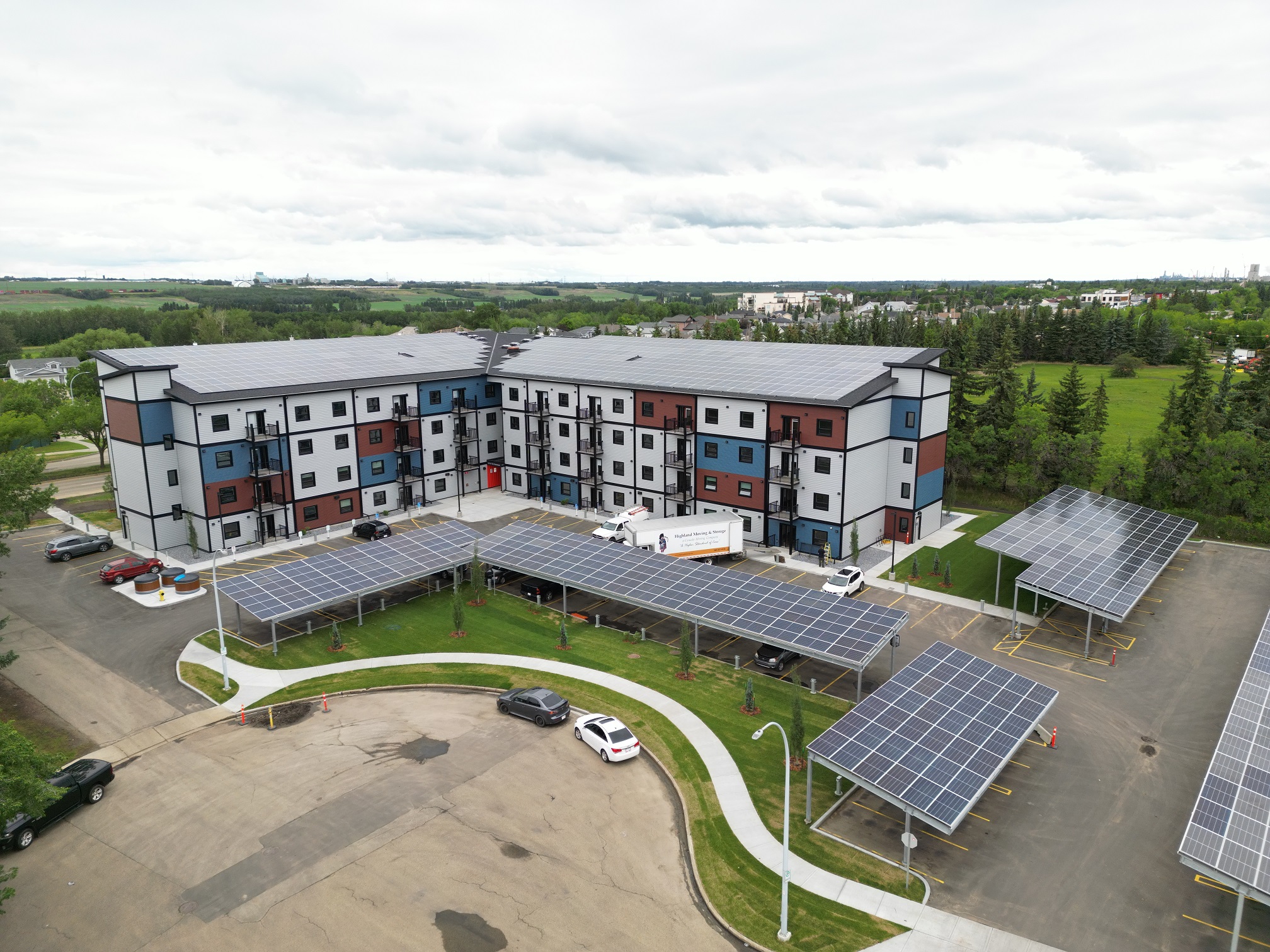 Vue aérienne grand angle du complexe de la Heartland Housing Foundation à Fort Saskatchewan, en Alberta. Il se compose de deux bâtiments contigus, de quatre étages chacun, dont l'extérieur est orné d'un motif de mosaïque bleu, rouge et blanc. Les toits sont recouverts de panneaux solaires, et le parking comporte également plusieurs grands panneaux solaires qui servent chacun de couverture au-dessus de 5 à 10 places de stationnement.