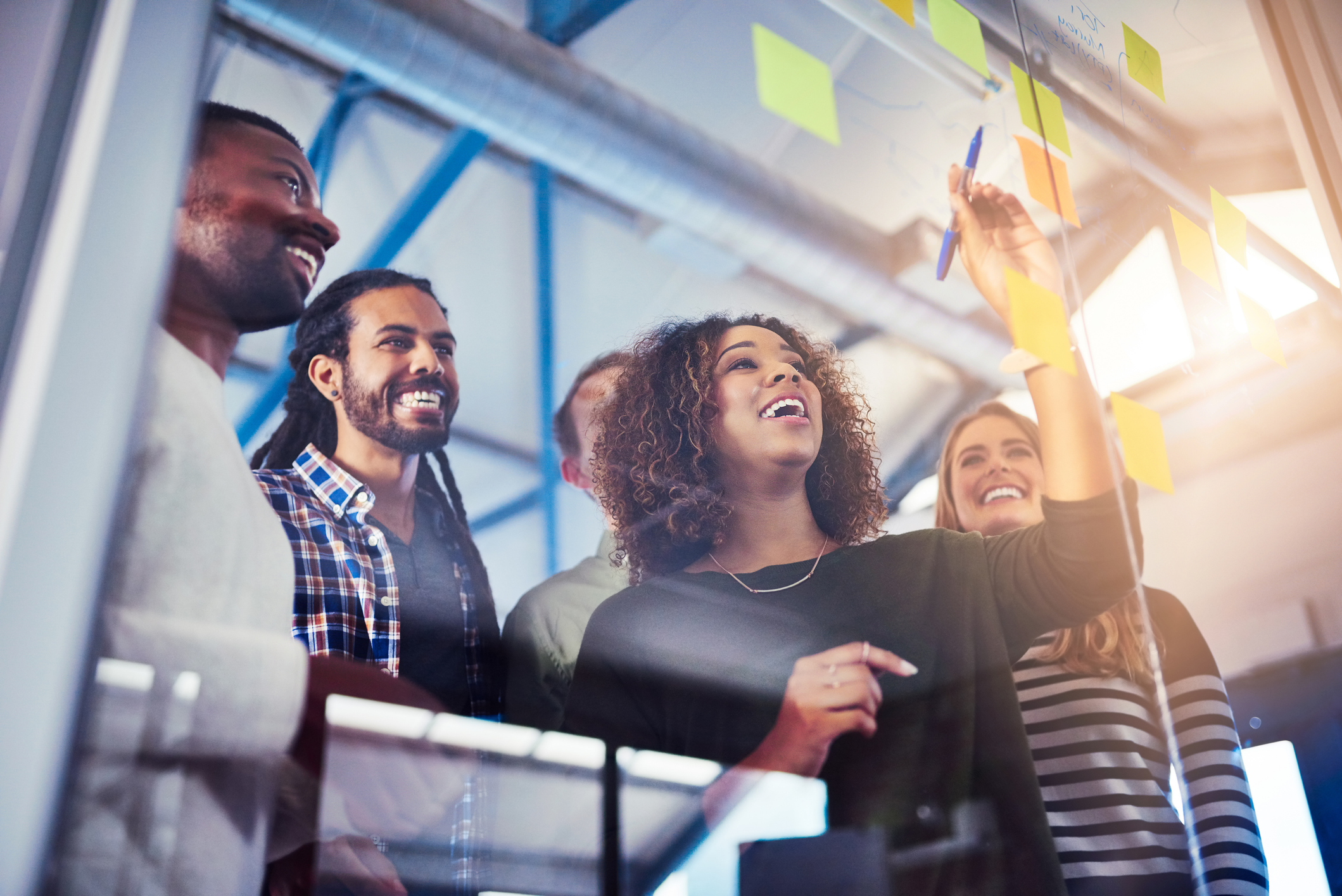 Diverse group of professionals smiling and collaborating in an office, with one person writing on a glass wall covered in sticky notes.