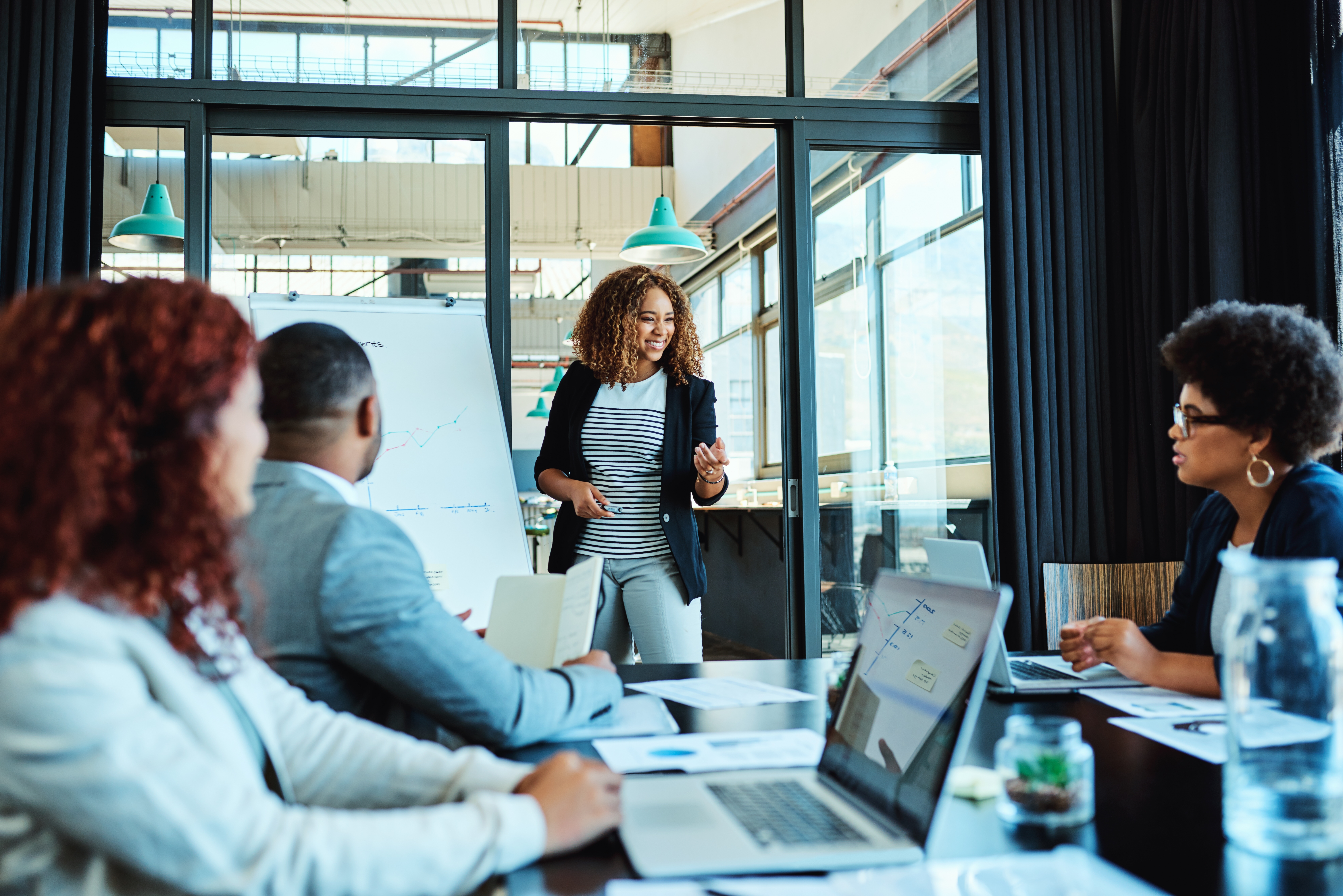 A woman stands by a whiteboard giving a presentation to a gathering of business colleagues at a boardroom desk