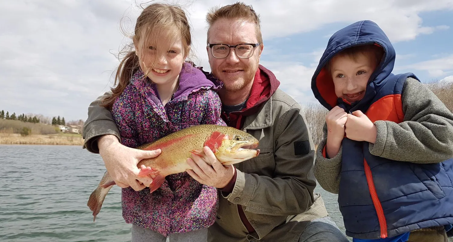 A man poses with two children, showing off a fish they caught in the trout pond.