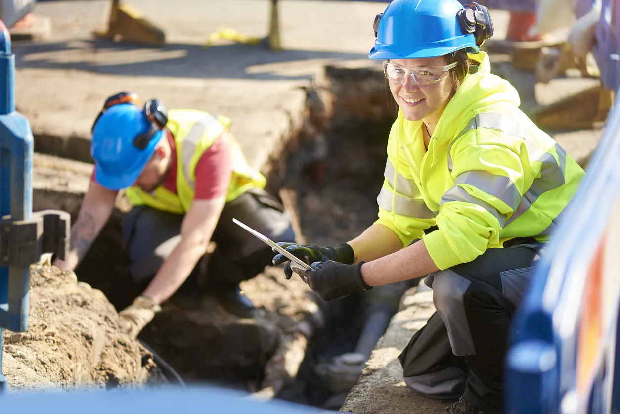 Female engineer kneeling outdoors in safety equipment inspecting a system of pipes used to transport energy.