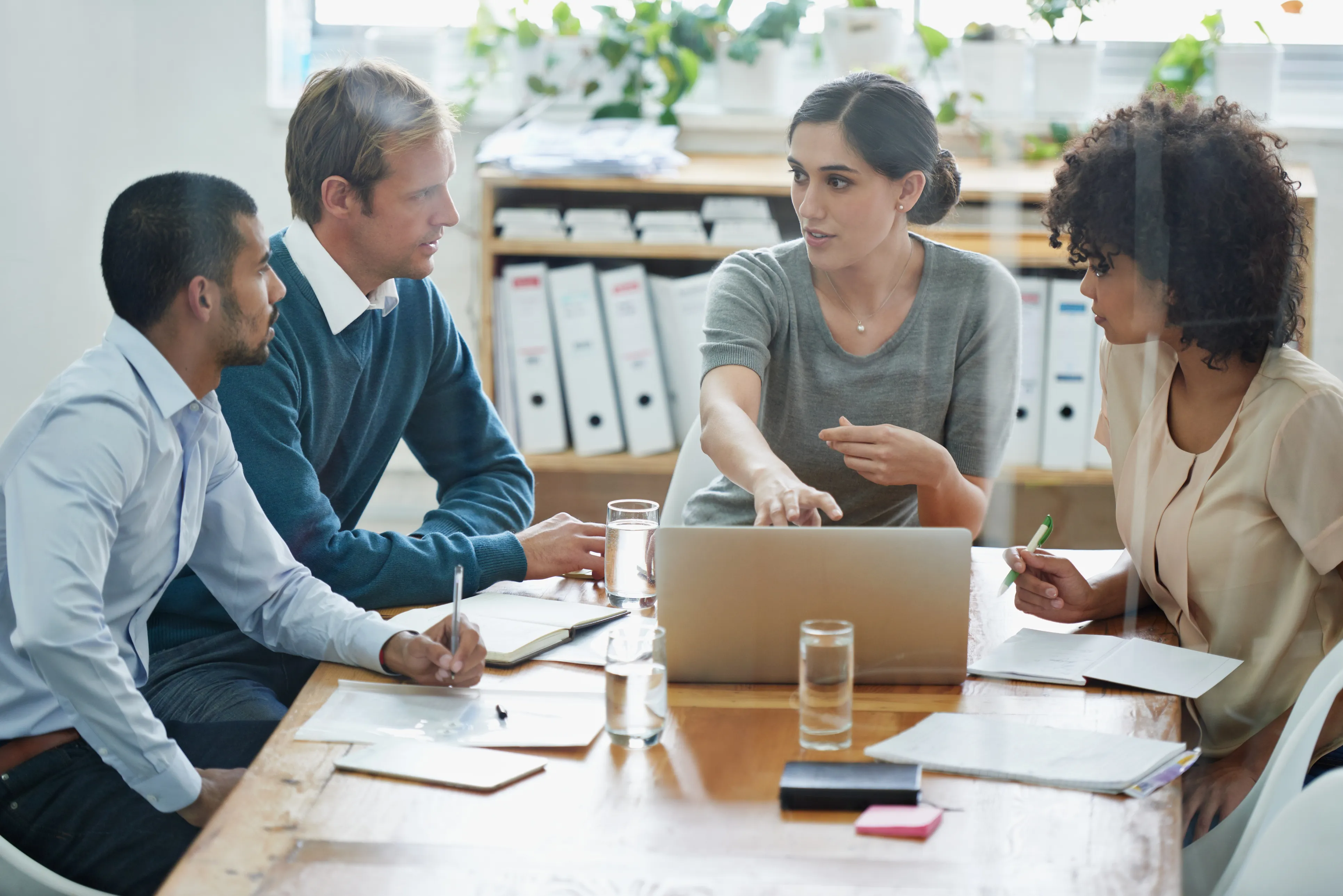 Four professionals in a meeting, engaged in discussion around a laptop. One woman is gesturing while others take notes, representing collaboration and training. 