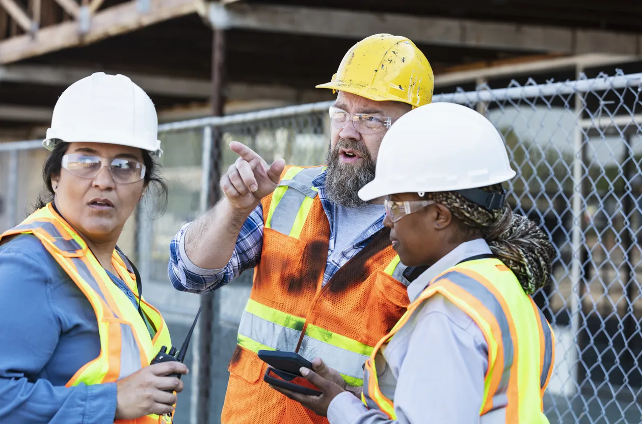 A diverse team of municipal workers in safety gear discusses project plans at an outdoor construction site.