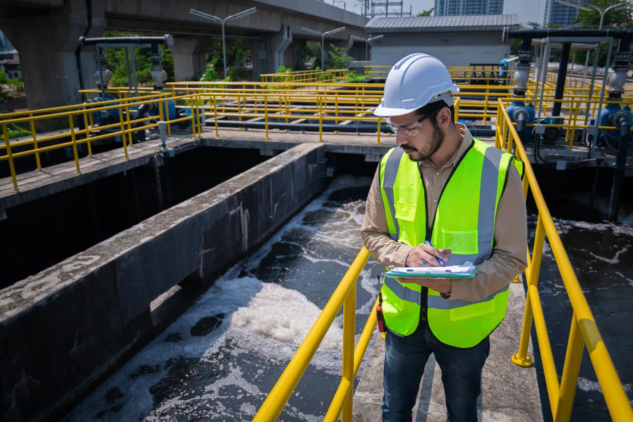 A municipal worker in a hard hat and safety vest inspects a water management facility, taking notes on a clipboard.