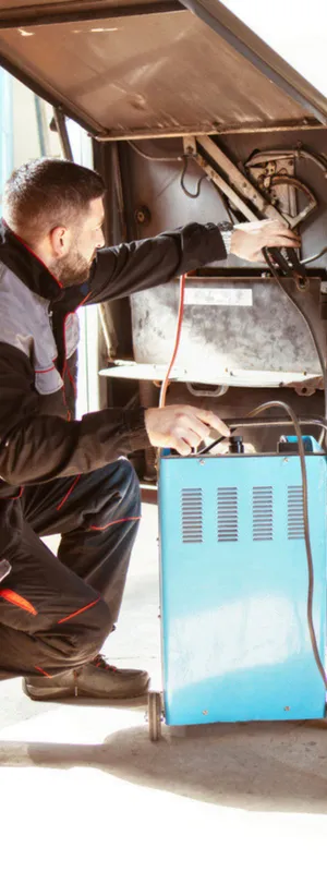 Man in a brightly lit garage kneels as he charges an electric bus with a battery charger. 