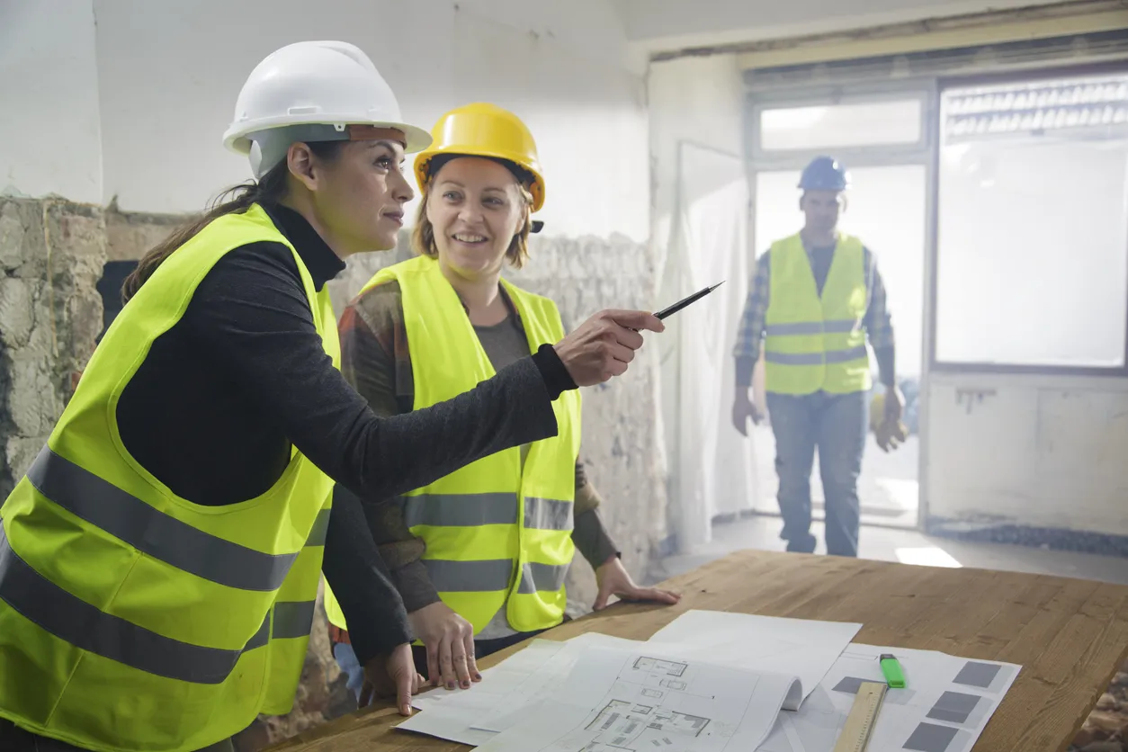Three construction workers in safety vests and hard hats look over plans in a bright sunny room.