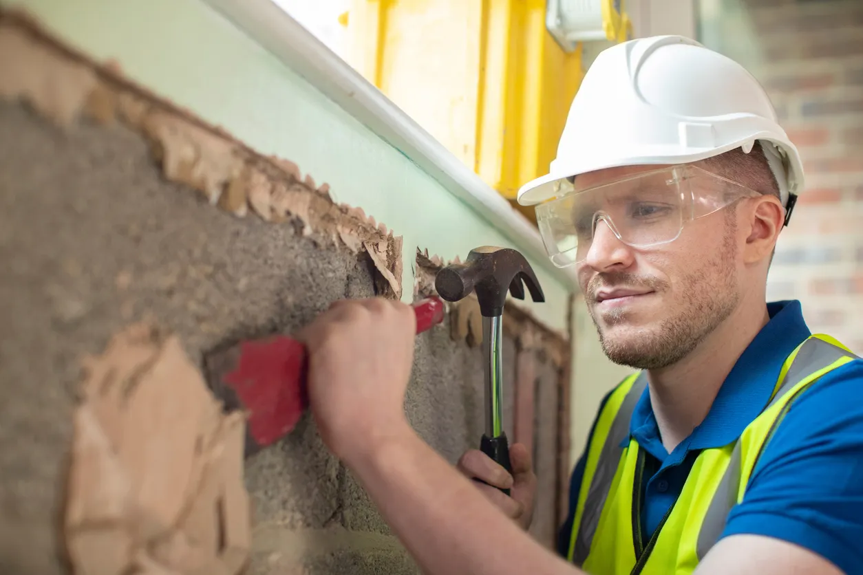 Worker wearing hardhat and safety glasses uses a chisel to remove plaster from wall.