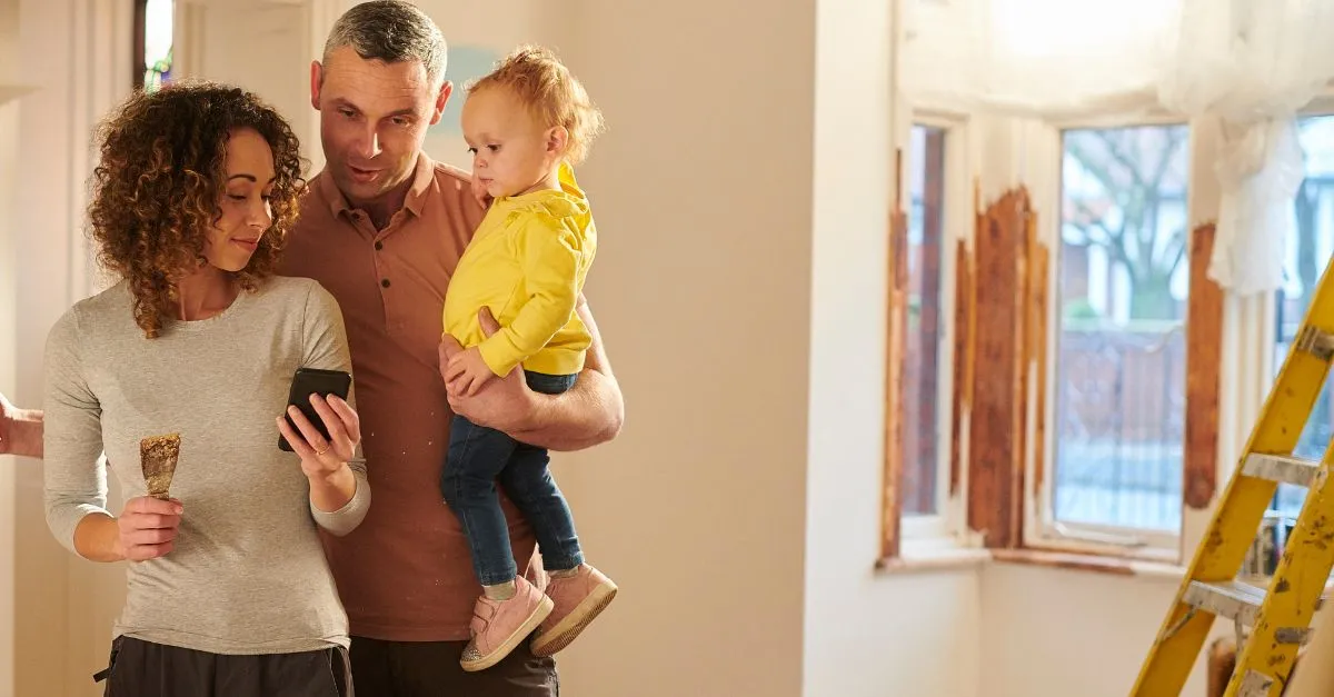 A woman shows something on her phone to her husband, who is standing next to her while holding their toddler in his arm. They stand in a home under renovation.